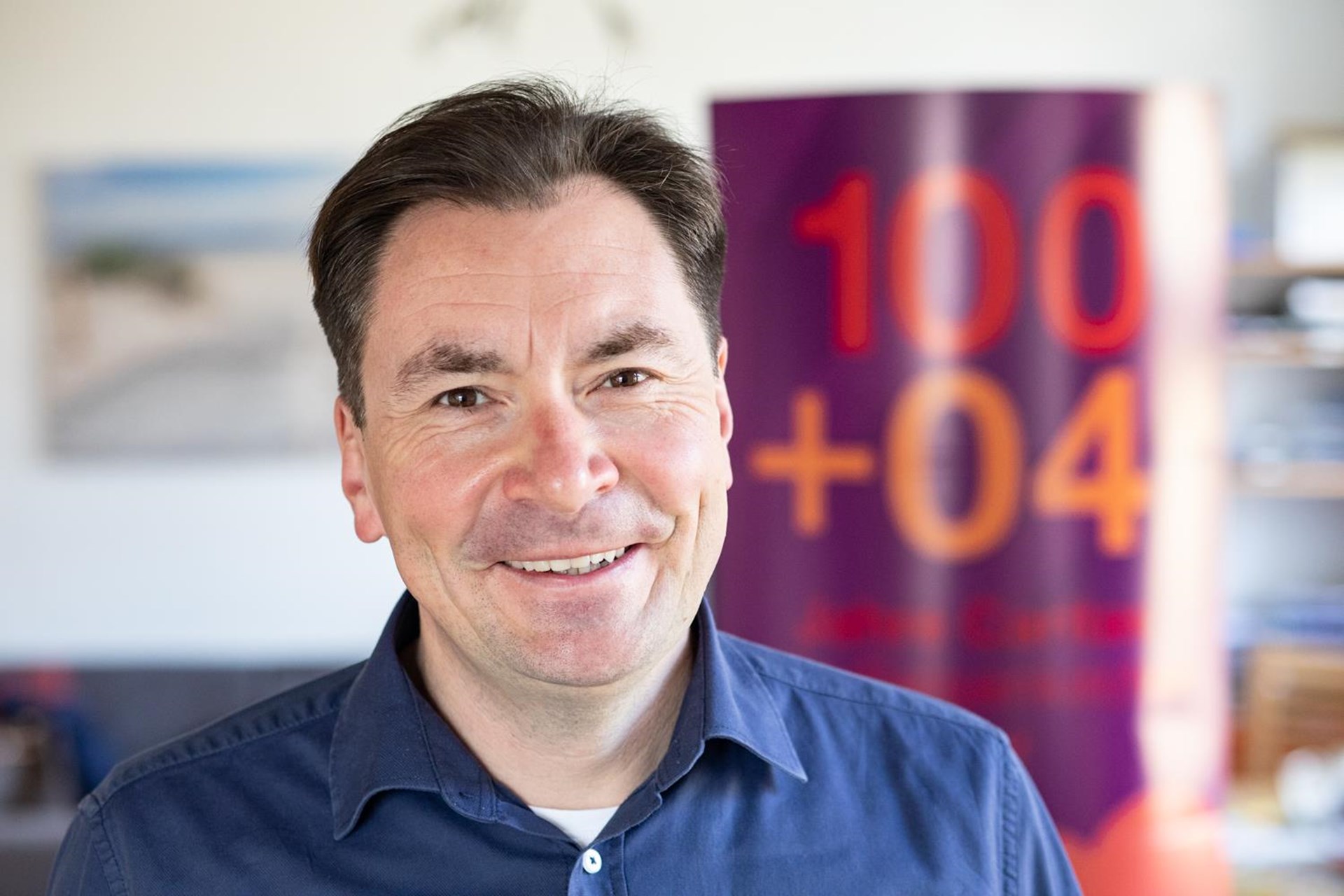 Eine Person steht in einem Innenraum und trägt ein dunkelblaues Hemd über einem weißen T‑Shirt. Im Hintergrund ist ein großer, violetter Banner mit orange‑roter Schrift „100 + 04“ zu sehen sowie ein unscharfer Raum mit Regalen und einem Wandbild.