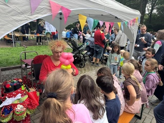 Mehrere Kinder stehen vor einem Stand unter einem Pavillon und beobachten eine Person beim Modellieren einer bunten Luftballonfigur. Im Hintergrund sind weitere Besucher:innen des Festes zu sehen.