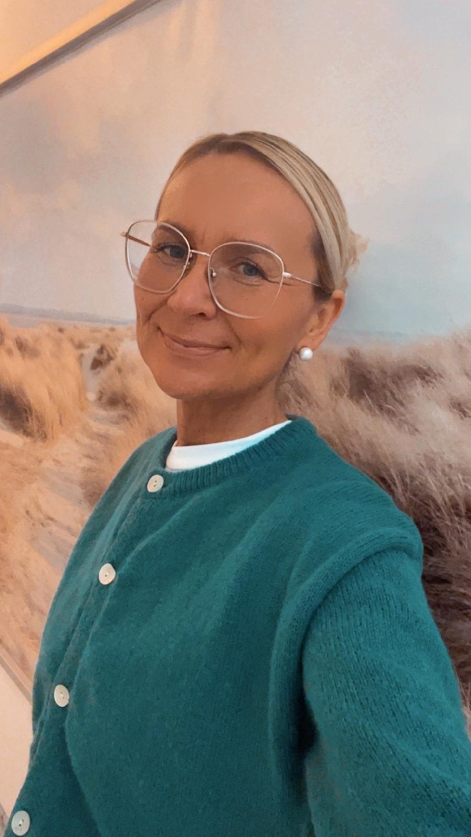 Eine Person steht vor einem großformatigen Wandbild, das eine Dünenlandschaft mit hellem Sand und trockenem Gras zeigt. Die Person trägt eine petrolfarbene Strickjacke mit weißen Knöpfen über einem weißen Oberteil und hat kurz geschnittenes, helles Haar.